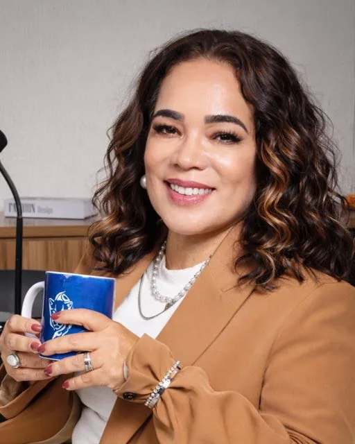 Paula Hernandez, palestrante e mentora em comportamento e vendas, sorrindo em seu escritório de blazer caramelo